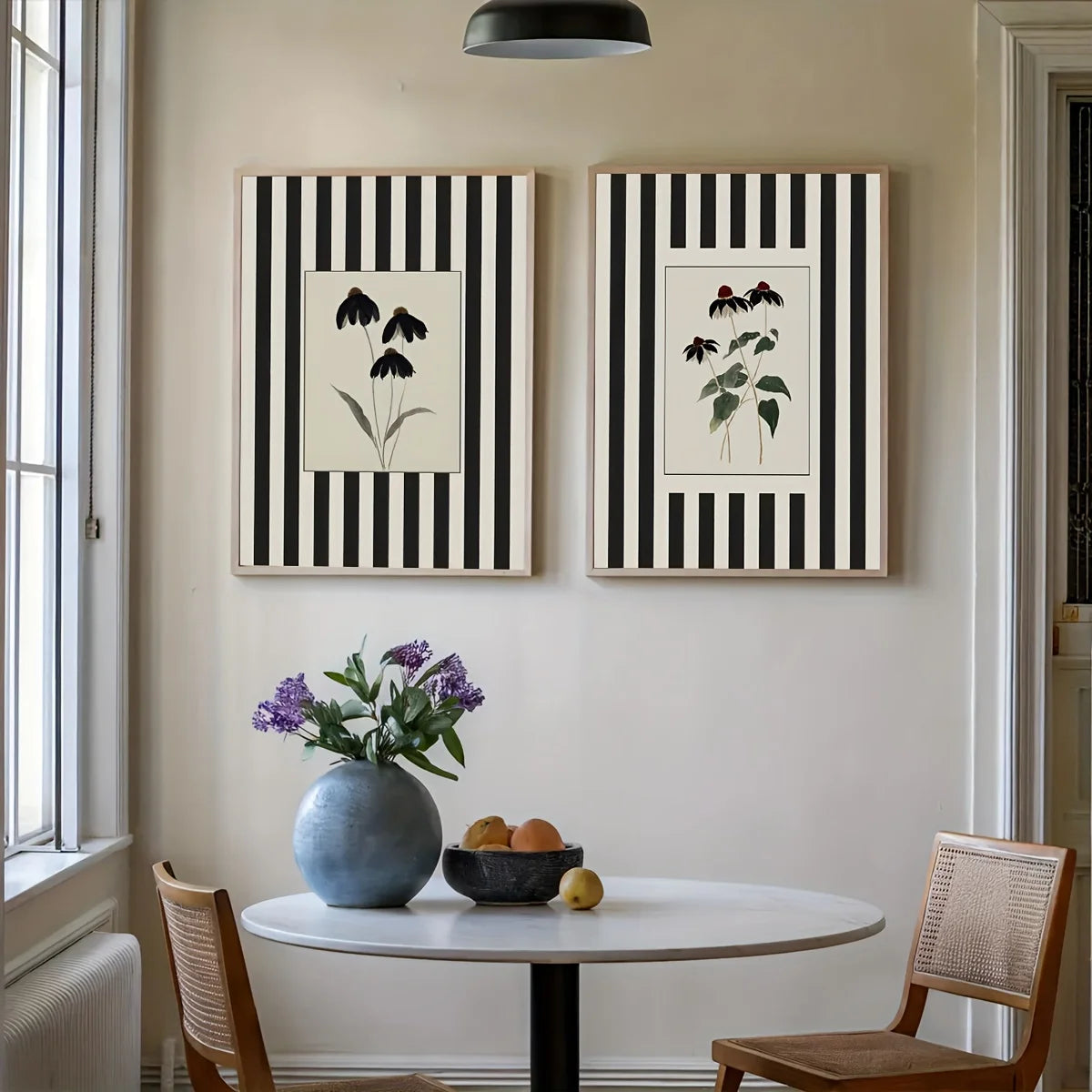 Two botanical art prints with black and white vertical stripes hang on a beige wall above a round dining table with chairs, a blue vase of purple flowers, and a bowl of fruit.