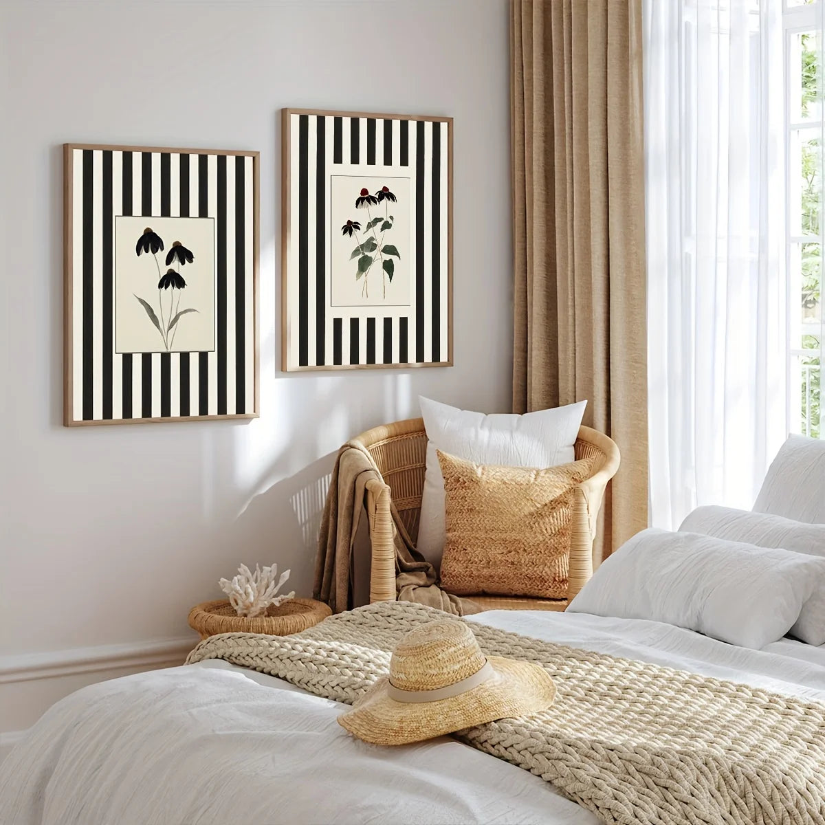 Two botanical art prints with black and white vertical stripes hang on a beige wall above a bed in a styled bedroom. The room includes a woven chair, neutral-toned bedding, a straw hat, and sheer curtains.