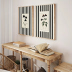 Two framed botanical art prints with black and white vertical stripes hang on a light wall above a wooden console table with books.