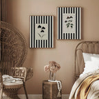 Two botanical prints with black and white vertical stripes hang on a beige wall in a styled bedroom. A wicker peacock chair, metal bed frame, small woven side table with a vase of dried flowers, and neutral bedding are visible.