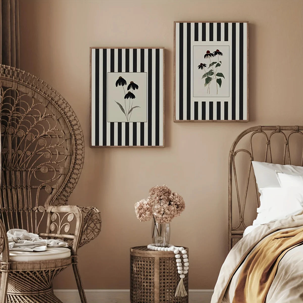 Two botanical prints with black and white vertical stripes hang on a beige wall in a styled bedroom. A wicker peacock chair, metal bed frame, small woven side table with a vase of dried flowers, and neutral bedding are visible.