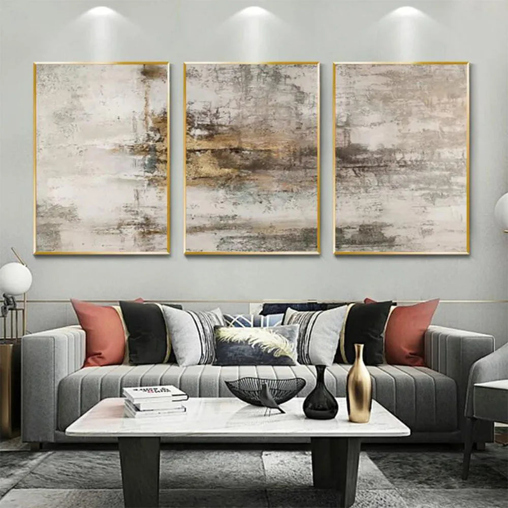 Three-piece abstract canvas wall art set with gold frames hanging above a gray sofa in a modern living room setting.