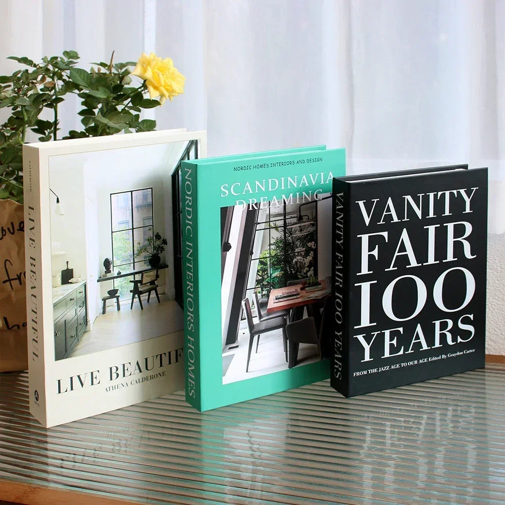 Three books titled 'Live Beautifully', 'Scandinavian Interiors Dreaming', and 'Vanity Fair 100 Years' on a reflective surface with a plant in the background.