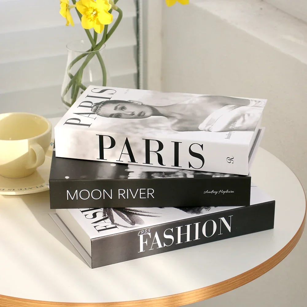 Stack of books titled 'Paris', 'Moon River', and 'Fashion' on a table with a cup and yellow flowers.