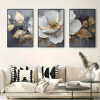 Three floral art pieces above a sofa with decorative pillows and a small table.