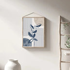 Framed abstract art with plant design on a wall next to a ladder and vase.