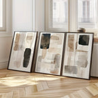 Three abstract art pieces in frames on a wooden floor with light streaming through windows.