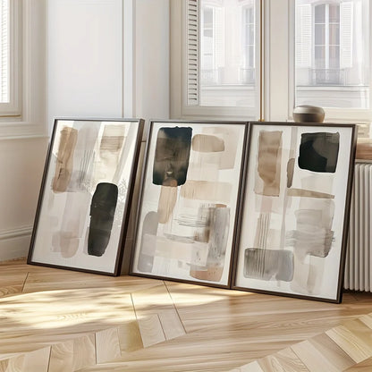 Three abstract art pieces in frames on a wooden floor with light streaming through windows.