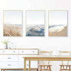 Three framed beach scenes on a wall above a dining table and chairs.