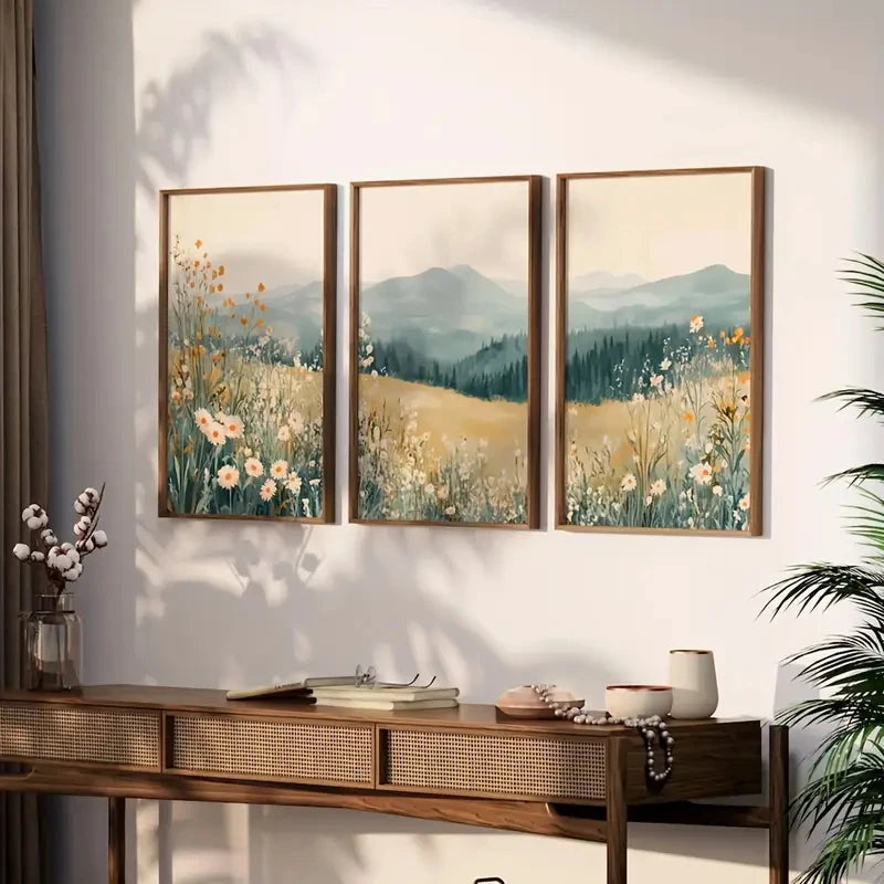 Three framed landscape paintings with mountains and flowers on a wall above a wooden console table.