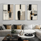 Three-piece abstract wall art set displayed above a modern sectional sofa in a living room setting. The minimalist canvases feature black, beige, gray, and white brushstrokes with subtle gold accents.