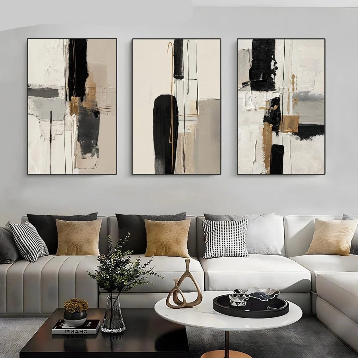 Three-piece abstract wall art set displayed above a modern sectional sofa in a living room setting. The minimalist canvases feature black, beige, gray, and white brushstrokes with subtle gold accents.