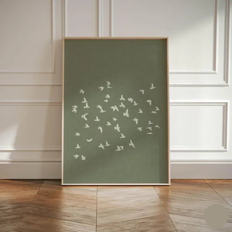 Framed artwork with white bird illustrations on a green background, displayed in a room with wooden flooring and white paneled walls.