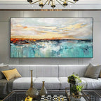A large abstract canvas painting hanging on a gray paneled wall above a modern light gray sofa in a styled living room. The artwork features a seascape-like composition with turquoise, teal, white, gray, and warm beige tones, plus soft orange and yellow highlights near the horizon.