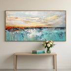 A large abstract landscape painting hangs on a light beige wall above a minimalist light wood console table. The painting features a vibrant turquoise and teal water scene with a warm orange and yellow sunset sky.