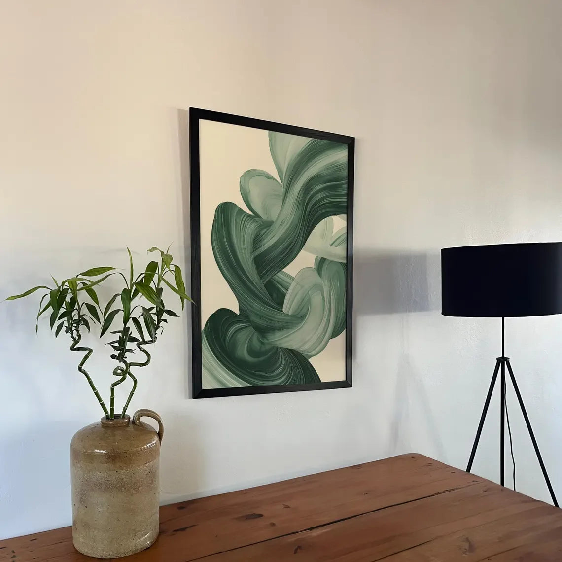 Framed abstract green artwork on a wall with a plant and lamp in the foreground.