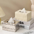 Two tissue boxes, one silver and one beige, on a surface with books in the background.