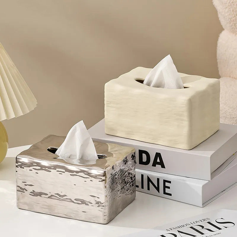 Two tissue boxes, one silver and one beige, on a surface with books in the background.