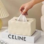 Beige tissue box with a hand pulling out a tissue, placed on a book titled 'Celine'.