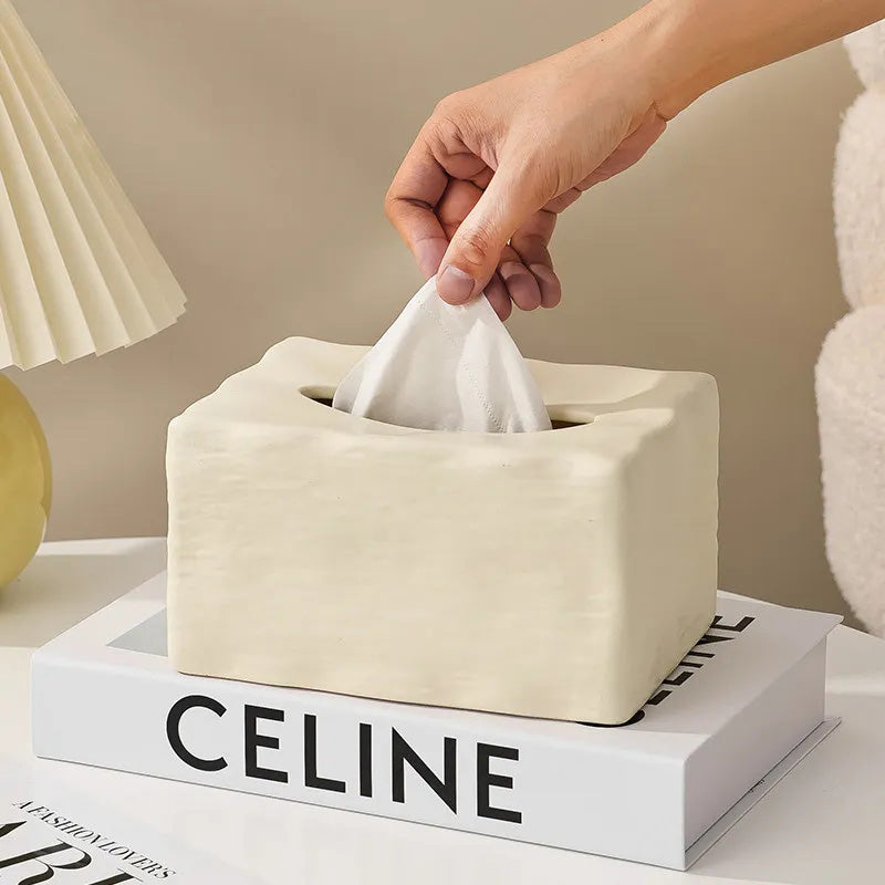 Beige tissue box with a hand pulling out a tissue, placed on a book titled 'Celine'.